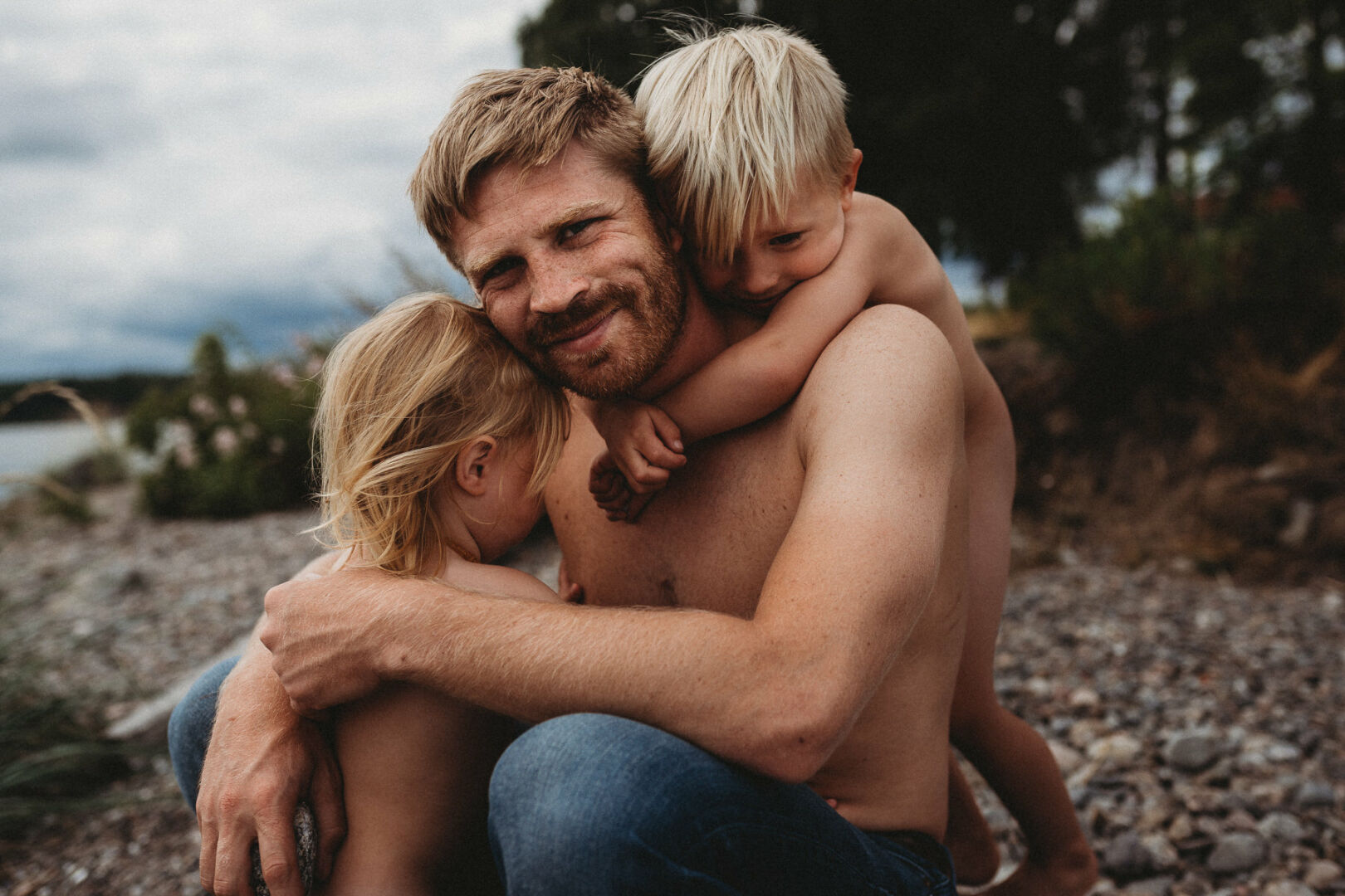 Dad getting hugged by his two children on the beach. Dad is looking in the camera, the kids are looking at dad.